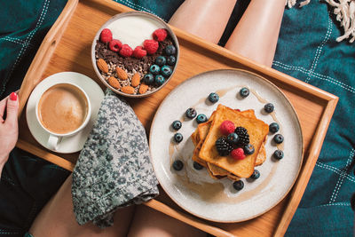 Breakfast in bed from above, yogurt bowl with berries, french toasts, coffee on plate, woman legs