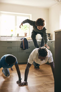 Playful son standing on father's back doing push-ups in kitchen at home