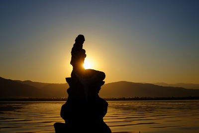 Silhouette statue by lake against sky during sunset