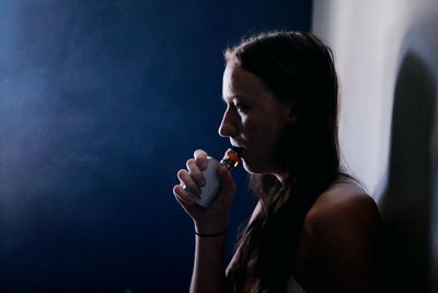Side view of young woman smoking electronic cigarette by wall at night
