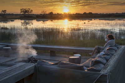 Woman drinking coffee on sofa against lake during sunset