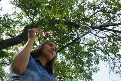 Low angle view of young woman blowing bubbles against tree