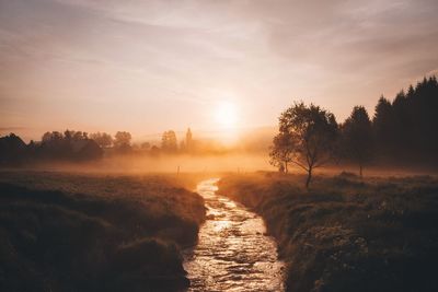 Stream flowing amidst field against sky during sunset