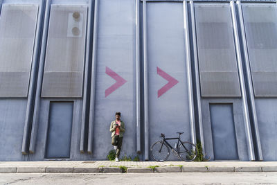 Man with smart phone leaning on factory wall near bicycle at footpath