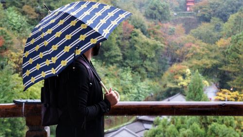 Side view of man carrying umbrella while standing by railing on mountain during rainy season