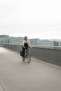Businesswoman cycling on footpath near railing against sky in city