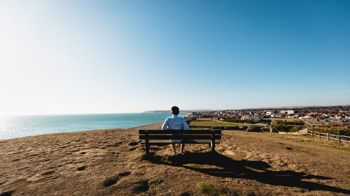 Rear view of man sitting on bench at beach against clear sky