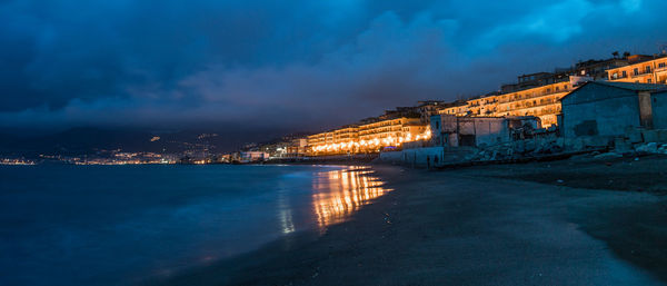 Illuminated buildings by sea against sky at night