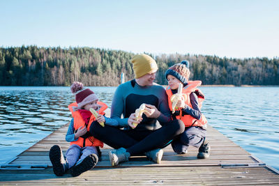 Dad sat on a jetty in a wetsuit eating fruit with his kids by a beach