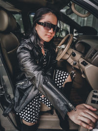 A girl wearing a polkadot dress, jacket, sun glasses and high boots sitting in a car 