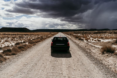 Spain, navarre, storm clouds over car driving along empty dirt road in bardenas reales