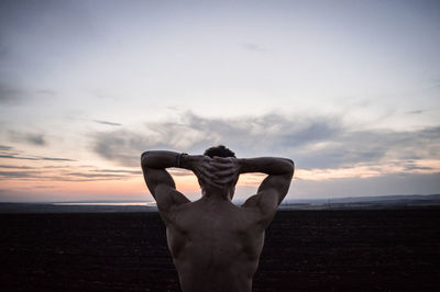 Rear view of shirtless man standing at beach with head in hands at dusk