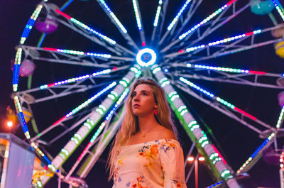 Low angle view of young woman standing against illuminated ferris wheel at amusement park
