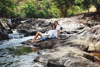 Portrait of smiling young couple sitting on rock by water