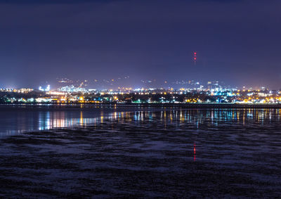 Illuminated buildings by river against sky at night