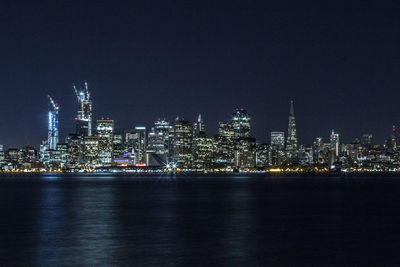 Illuminated cityscape by sea against clear sky at night