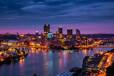 Illuminated buildings by river against sky in city at dusk