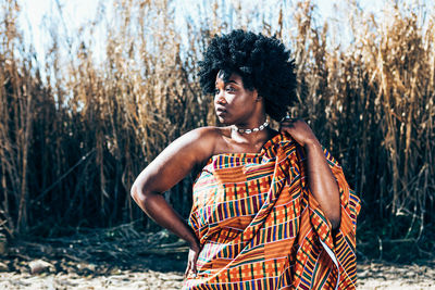 Serious african model with afro hairstyle wearing colorful handwoven kente and looking away while standing on field on bright summer day