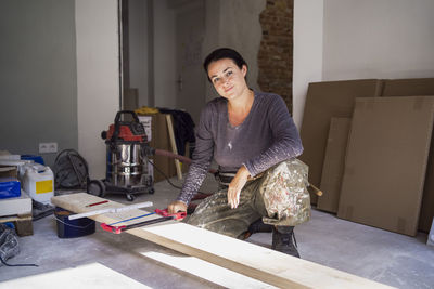 Portrait of confident female carpenter holding handsaw crouching in front of plank in apartment