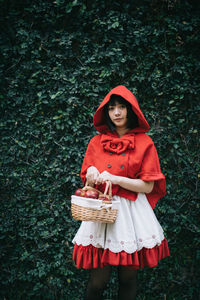 Woman in little red riding hood costume holding basket while standing against plants