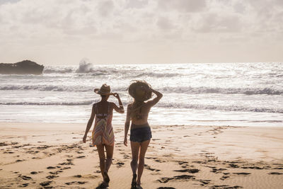 Rear view of women wearing hats while walking at beach towards seascape during sunset