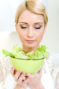 Beautiful young woman holding fresh salad bowl with eyes closed at home