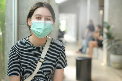 Teen woman wearing medical mask looks at camera, european teenager wearing t-shirt sitting in cafe.