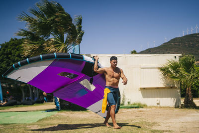 Young shirtless man carrying wing foil at beach on sunny day