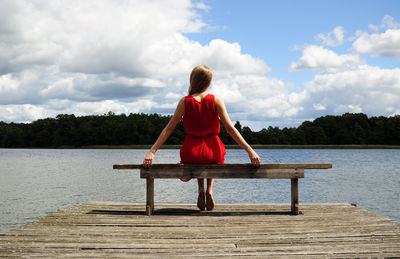 Rear view of woman wearing red dress sitting on bench at jetty over lake against sky