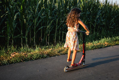 Girl in flower dress riding red scooter on a country road next to corn field