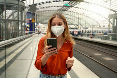 Portrait of young woman with ffp2 protective face mask using smartphone in train station on sunset