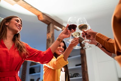 From bellow side view of cheerful diverse friends celebrating and clinking toasting with wineglasses while standing at served table with delicious dinner at home