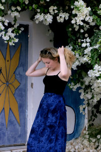 Beautiful woman with hair in hands standing against wall and flowering plants