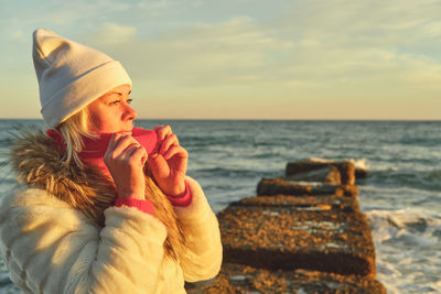 A woman with blond hair in a hat and a fur coat walks along the sea along a stone breakwater