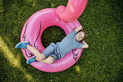 Blond boy in blue clothes, lying on pink inflatable flamingo on grass.