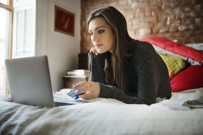 Woman shopping online on laptop while lying on bed at home