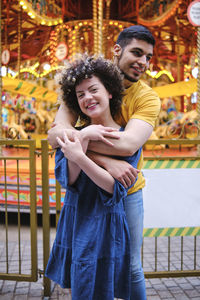 Happy young couple standing with arms around at amusement park