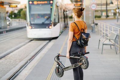 Back view of woman with backpack and folded electric scooter waiting at tram stop