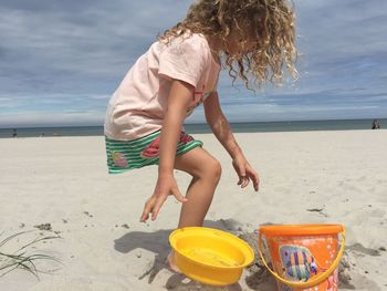 Side view of girl throwing plastic containers on sand at beach against sky