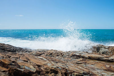 Waves splashing on rocks at shore against blue sky