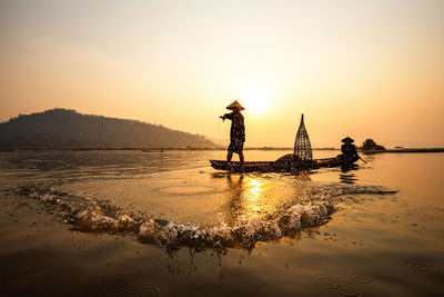 Silhouette of people fishing in boat on lake against sky during sunset