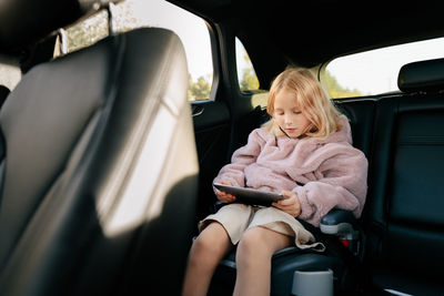 Girl with blond hair looking at camera while sitting in passenger seat of automobile with modern tablet during ride