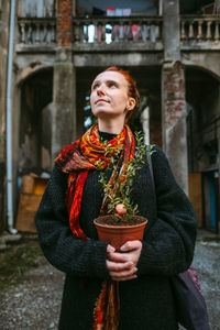 Thoughtful woman holding potted plant while standing against building