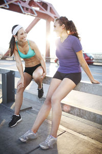 Happy friends talking while sitting on railing at bridge during sunny day