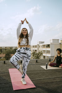 Full length of smiling young woman practicing tree pose by friend practicing breathing exercise on rooftop