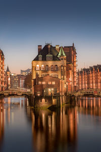 Illuminated buildings by river against sky in city at dusk