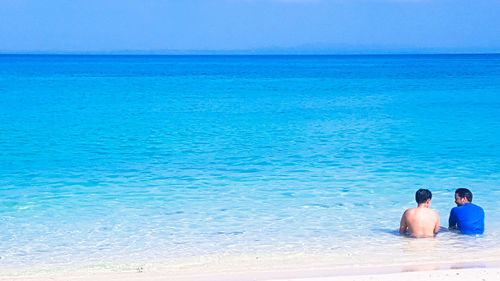 Rear view of men relaxing at beach against clear blue sky