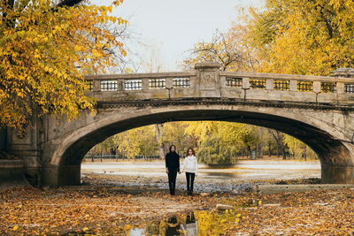 Lesbian couple holding hands while standing at arch bridge during autumn