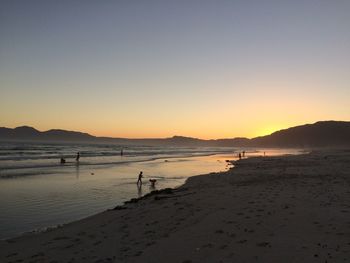 People enjoying at beach against clear sky during sunset