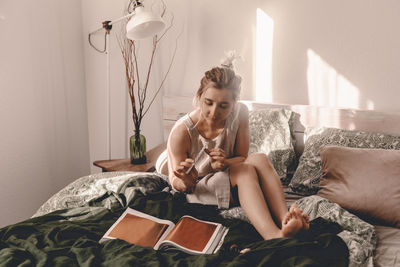 Full length of woman reading book while sitting on bed at home